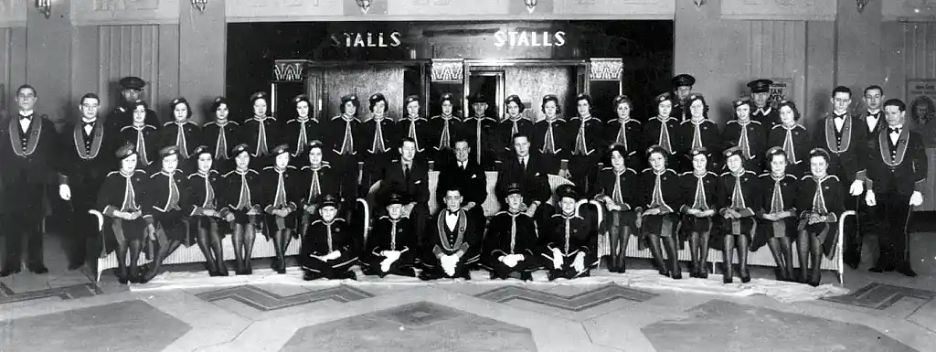 Formal 1940s group portrait of a large group of uniformed cinema staff, sitting and standing in a semi-circle in the foyer of a cinema.