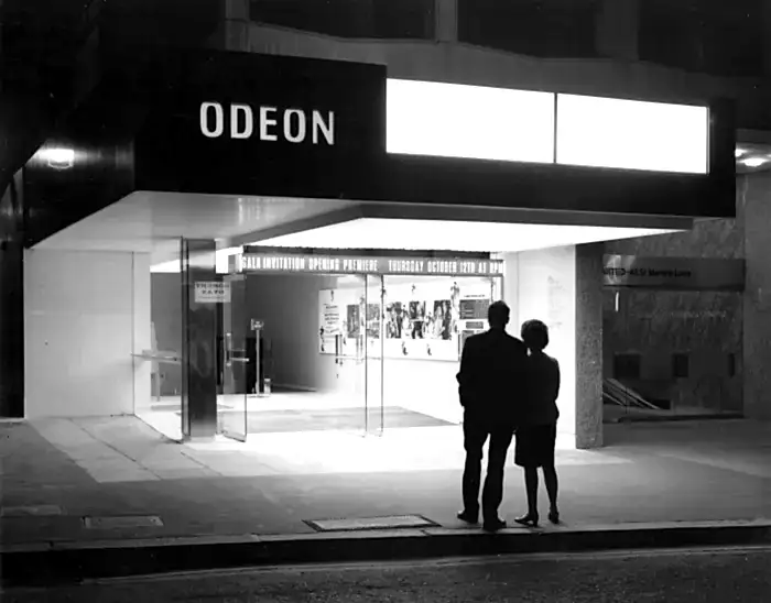 A black and white photo of a silhouetted couple standing with their backs to the camera in front of the entrance to Odeon cinema. A sign above the doors advertises a 'gala invitation opening premiere'.