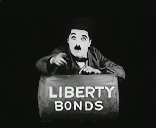 Chaplin's Little Tramp character looking into camera with a beseeching look while leaning on a barrel marked 'Liberty Bonds'.
