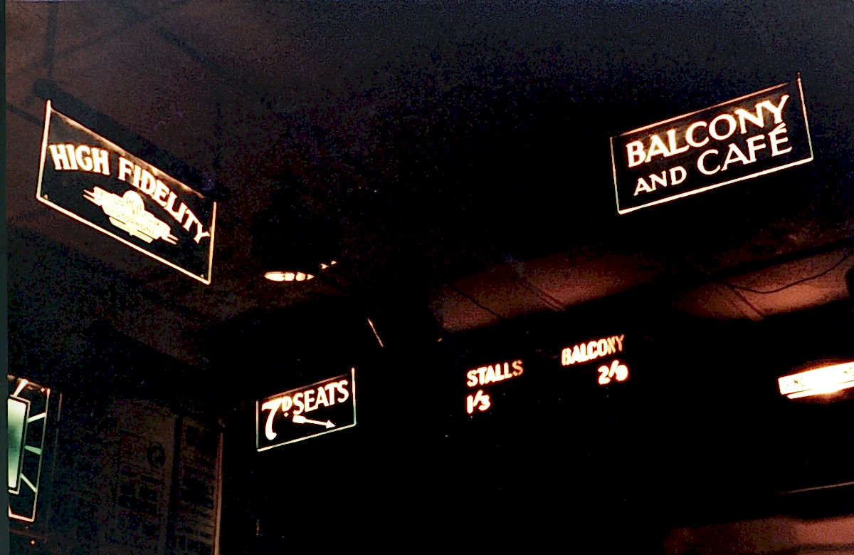 A group of illuminated glass signs shown against the dark background of a cinema lobby. One reads "High Fidelity", the others direct patrons to seating areas and the cafe.